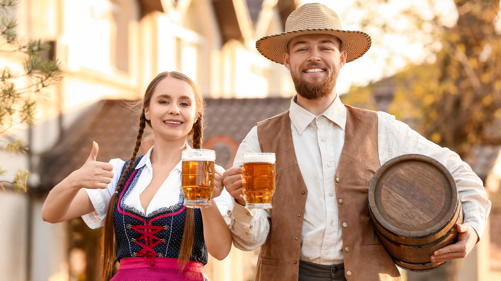 Woman and man with hat both wear traditional German clothing. The woman holds her right hand, thumb up and her left hand holds a stein of beer. The man has a small barrel under his left arm and his right hand holds a stein of beer. The blured background is a German village.