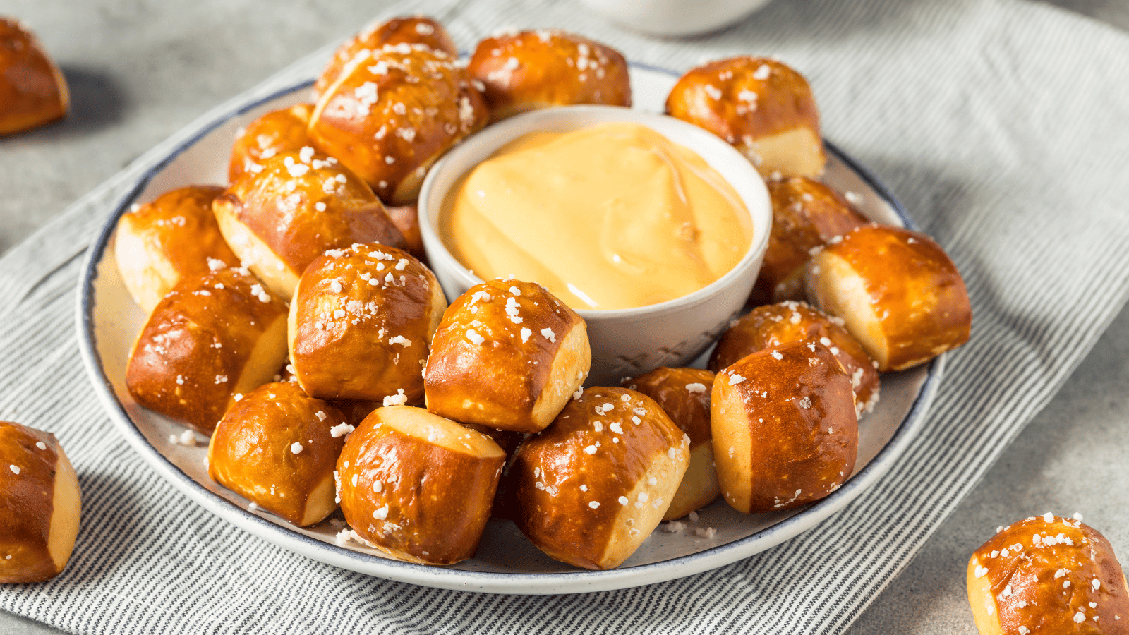 White plate with skinny blue line on top sits on a stripped napkin. One the plate is a pile of pretzel bites and some beer cheese dip in a white ramekin. Scattered around the plate are a few extra pretzel bites
